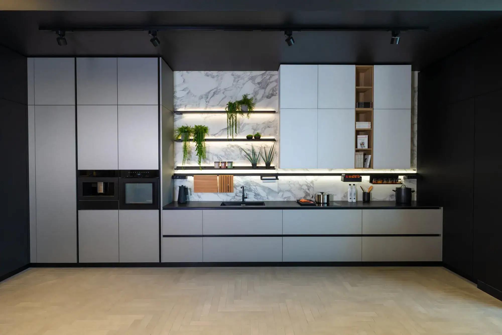 A Framework kitchen wall with tall grey units, built-in ovens, marble backsplash and lit shelves